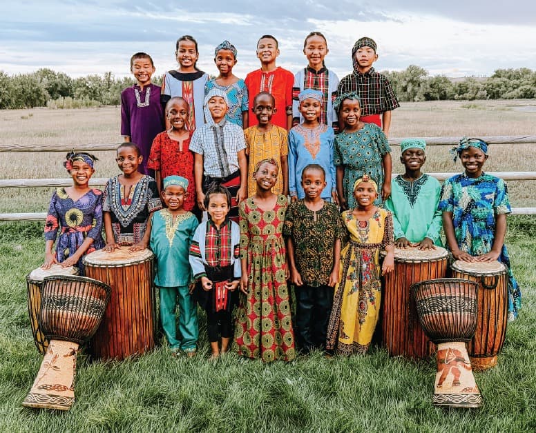 The 2026-2027 His Little Feet International Children's Choir in colourful traditional dress, standing in a field behind djembe drums
