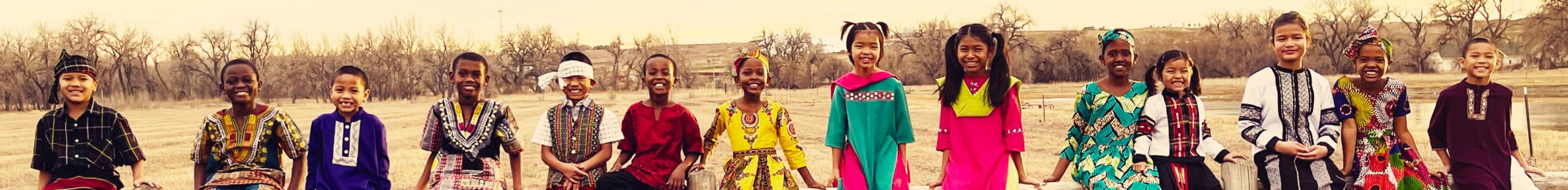 Fifteen choir children in colourful traditional dress sitting in a row on a fence, smiling